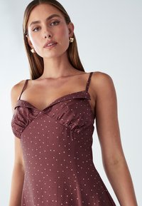 Hnedé šaty s bodkami, s priliehavým živôtikom, volánovým výstrihom a tenkými ramienkami. Hladká textúra látky s rozšíreným dizajnom sukne.