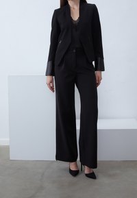 Černý pánský blejzr s koženými detaily na rukávech, spárovaný s černými kalhotami s vysokým pasem a černými špičatými lodičkami. Hladká textilie.