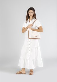 Kabešová taška béžové barvy s hnědými akcenty, na které se nachází opakující se vzor loga. Modelka má na sobě bílé košilové šaty a hnědé sandály.