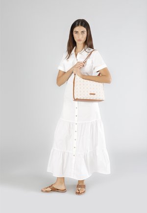 Beige schoudertas met bruine accenten, voorzien van een herhalend logo-patroon. Model draagt een witte blousejurk en bruine sandalen.