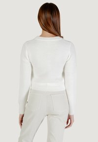 Donna con i capelli castani lisci sta rivolta di spalle, indossando un maglione bianco morbido e pantaloni avorio con tasche posteriori.