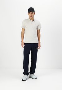 Beige Polo-Shirt mit Logo auf den Ärmeln, marineblaue Hose mit Logo-Akzent und grauen Sneakers. Das Model trägt eine schwarze Mütze und steht vor einem weißen Hintergrund.