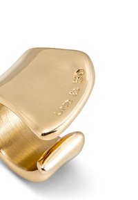 Anillo de metal en tono dorado con un diseño liso y curvado, que presenta una superficie pulida y marcas en relieve en un lado.
