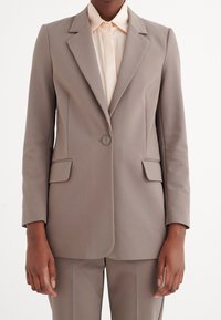 Blazer color beige con un design su misura, due tasche frontali, chiusura a un bottone e rever a snotch, abbinato a una camicia beige.
