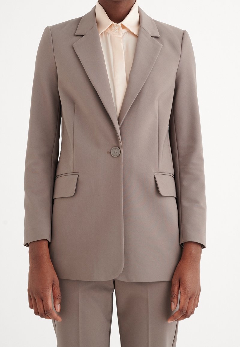 Blazer color beige con un design su misura, due tasche frontali, chiusura a un bottone e rever a snotch, abbinato a una camicia beige.