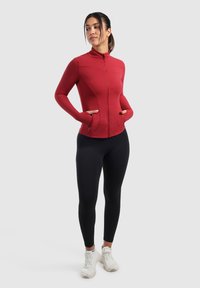 Rød zip-up sportsjakke med lange ærmer, der har en høj krave og sidelommer, som er sat sammen med sorte leggings og hvide sneakers.