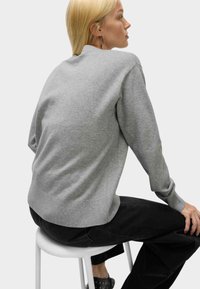 Szary sweter dziany z okrągłym dekoltem, ściągaczami przy mankietach i u dołu; noszony z czarnymi spodniami o gładkiej fakturze, siedzący na białym stołku.