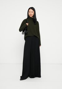 Maglione oversize di un verde scuro e pantaloni neri a gamba larga plissettati. Il maglione ha un collo alto; i pantaloni hanno un aspetto fluido e texturizzato.