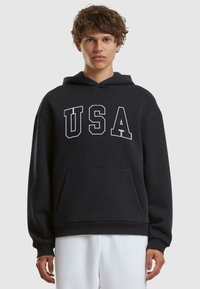 Sudadera con capucha negra con "USA" en letras blancas en negrita, con un bolsillo frontal y puños y dobladillo acanalados. Textura de tela suave.