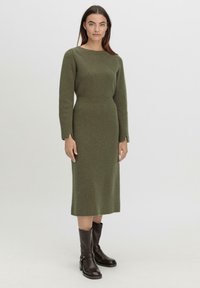Langes, enganliegendes Kleid aus ribbigem, olivgrünem Strickmaterial. Verfügt über einen runden Ausschnitt und lange Ärmel. Kombiniert mit braunen Leder-Stiefeletten.