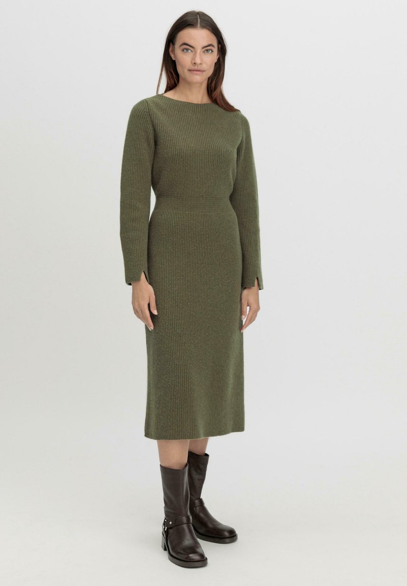 Langes, enganliegendes Kleid aus ribbigem, olivgrünem Strickmaterial. Verfügt über einen runden Ausschnitt und lange Ärmel. Kombiniert mit braunen Leder-Stiefeletten.