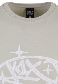 Felpa grigia chiara con scollatura a costine. Presenta un design grafico ricamato bianco con stelle e testo stilizzato. Etichetta di taglia: L.