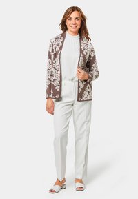 Cardigan à motifs marron et blanc porté sur un blouse blanche, associé à un pantalon blanc et des sandales à glisser blanches. Textures lisses, design ouvert.