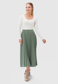 Długa zielona spódnica plisowana z wysokim stanem, połączona z białą bluzką z długim rękawem, z falbanym dekoltem i gładką fakturą.
