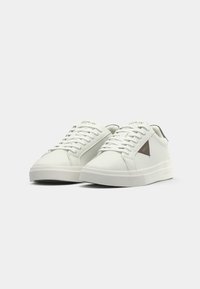 Pair of white low-top sneakers with white laces and a small black and red triangular logo on the sides on a plain background.