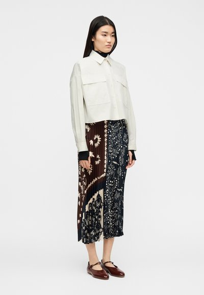 Γυναίκ α που φοράει λευκό crop top, πλισέ midi φούστα με καφέ και ναυτικά μοτίβα, και σκουρόκόκκινα παπούτσια Mary Jane, στέκεται μπροστά σε λευκό φόντο.