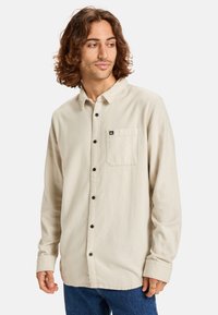 Light beige long-sleeve shirt with a button-down collar, front pocket, and brown buttons; soft texture and relaxed fit.