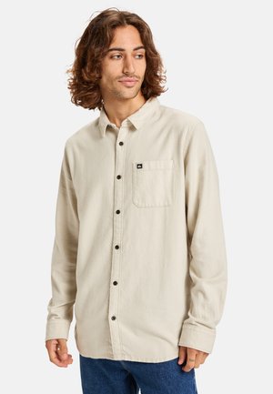 Light beige long-sleeve shirt with a button-down collar, front pocket, and brown buttons; soft texture and relaxed fit.