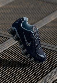 Melanīgā un gaiši zilā Nike Shox skriešanas apavi ar gaisa spilvenu zoli, kas atpūšas uz metāla režģa virsmas.