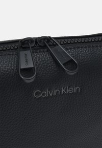 Borsa in pelle nera con superficie testurizzata, caratterizzata da doppi tiranti neri per la cerniera e logo "Calvin Klein" impresso sul davanti.