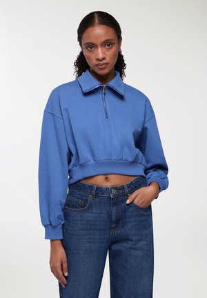 Felpa blu cropped con colletto alto e chiusura a mezza zip, abbinata a jeans in denim blu scuro con tasche classiche.