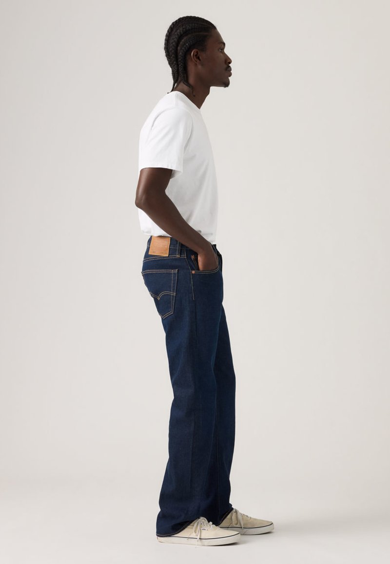 Blue denim jeans with a straight cut, featuring a brown leather patch on the back, and side pockets. Worn with a white t-shirt and sneakers.
