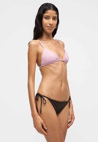 Femme aux longs cheveux foncés portant un haut de bikini rose clair et un bas de bikini noir avec des liens sur les côtés, se tenant devant un fond clair uni.