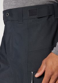 Zwarte, waterdichte broek met een riem, verstelbare klittenbandsluiting, zijzak met rits en een gladde textuur.
