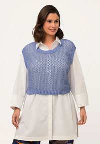 Ljusblå stickad cropped top med bred halsringning och ärmlös design, bärs över en vit skjorta med knappar. Strukturerat mönster synligt.