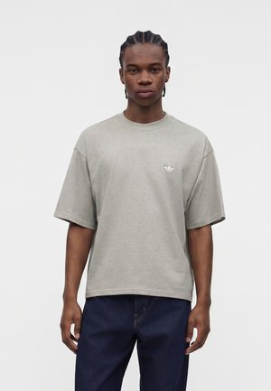 Ung mand med flettet hår iført en løs, lysegrå Adidas t-shirt og mørkeblå jeans, stående foran en ensfarvet hvid baggrund.