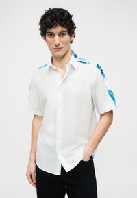 Νέος άνδρας με σγουρά μαλλιά, φορώντας λευκό πουκάμισο με κοντά μανίκια και μπλε σχέδια στους ώμους, καθώς και μαύρο παντελόνι, στέκεται με το ένα χέρι στην τσέπη.