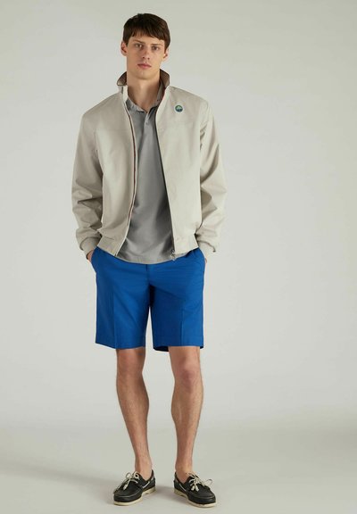 Licht beige ritsjack met een contrasterende bruine binnenkant, grijs poloshirt en blauwe shorts. Casual schoenen maken de outfit compleet.