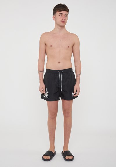 Pantaloni da bagno neri con elastico in vita e cordino bianco, con un design stampato sulla gamba sinistra. Indossati con sandali neri.