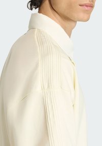 Koszula w kolorze kremowym z kołnierzykiem, szczegółami prążków na ramionach, gładką teksturą, luźnym krojem i subtelnym połyskiem. Zbliżenie z boku.
