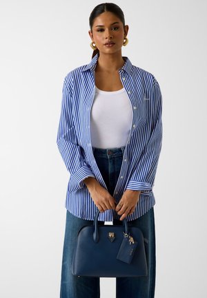 Donna con camicia a righe blu e bianche sopra una maglietta bianca, jeans blu, che tiene una borsa blu navy con dettagli dorati su sfondo semplice.
