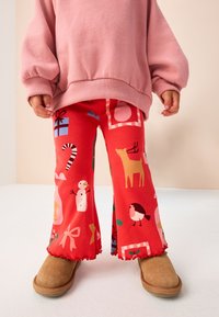 Pantalon évasé en velours côtelé rouge avec des motifs sur le thème des fêtes, incluant des rennes, des bonhommes de neige et des cadeaux, assorti à des bottines tan.