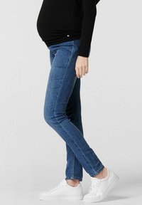 Modré těhotenské džíny se slim fit střihem, středním pasem a hladkou denimovou texturou. Kombinované s bílými teniskami. Klasický pětikapsový design.