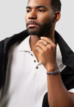 Silver ring on hand, white polo with dark buttons, navy bracelet, and a black shawl draped over shoulder against a grey background.