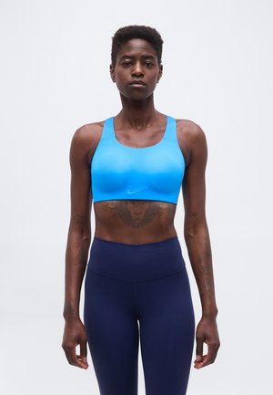 Atletisk kvinde iklædt en lyseblå sports-bh og mørkeblå leggings står med fronten mod kameraet foran en ensfarvet baggrund.