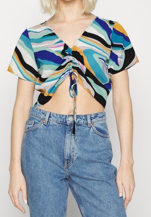 Kobieta w niebieskich, wysokich jeansach i krótki rękaw, kolorowy marszczony krótki top z dekoltem w serek i marszczonym wiązaniem w środku.