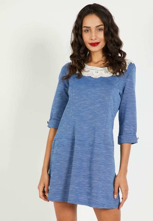 Vestido azul con mangas tres cuartos, presenta un cuello de encaje blanco, tejido texturizado y bolsillos frontales. Diseño simple y ajustado.