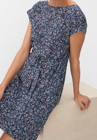 Marinblå klänning med kort ärm och blommönster i rosa och vitt, med en draperad midja och avslappnad passform. Mjuk textil och slät yta.