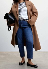 Vrouw in een camelkleurige jas, gestreepte shirt, blauwe jeans, zwarte ballerina's en met een zwarte handtas, tegen een effen achtergrond.
