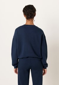 Tamsiai mėlynas džemperis laisvo silueto, su ribuojamomis rankogaliais ir juosmeniu, pasižymintis paprasta, lygią tekstūrą be matomų logotipų ar raštų.