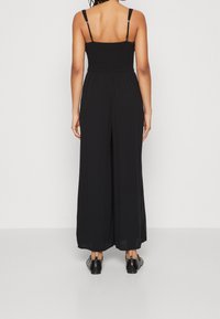 Kvinna med flätat hår, klädd i en ärmlös svart jumpsuit och svarta klänningesskor, stående vänd mot en vit bakgrund.