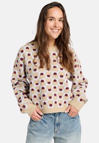 Gebreide trui in crème met een geruit patroon en rode accenten. Ontspannen pasvorm met geribde manchetten en zoom, gecombineerd met lichtblauwe jeans.
