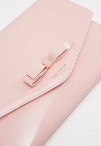 Pochette en cuir verni rose avec un détail en nœud, une quincaillerie dorée et le nom de la marque embossé. Texture lisse et design élégant avec des bords arrondis.