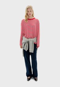 Rood-witte gestreepte langemouwen shirt, grijze trui om de taille geknoopt, gecombineerd met donkerblauwe flare jeans en zwarte puntige schoenen.