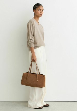 Femeie cu părul împletit, purtând un pulover bej și pantaloni albi largi, ținând o geantă mare maro, stând în fața unui perete alb.