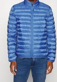 Gepolsterte blaue Jacke mit hohem Kragen und Frontreißverschluss, mit glänzender Textur und horizontalem Steppmuster.